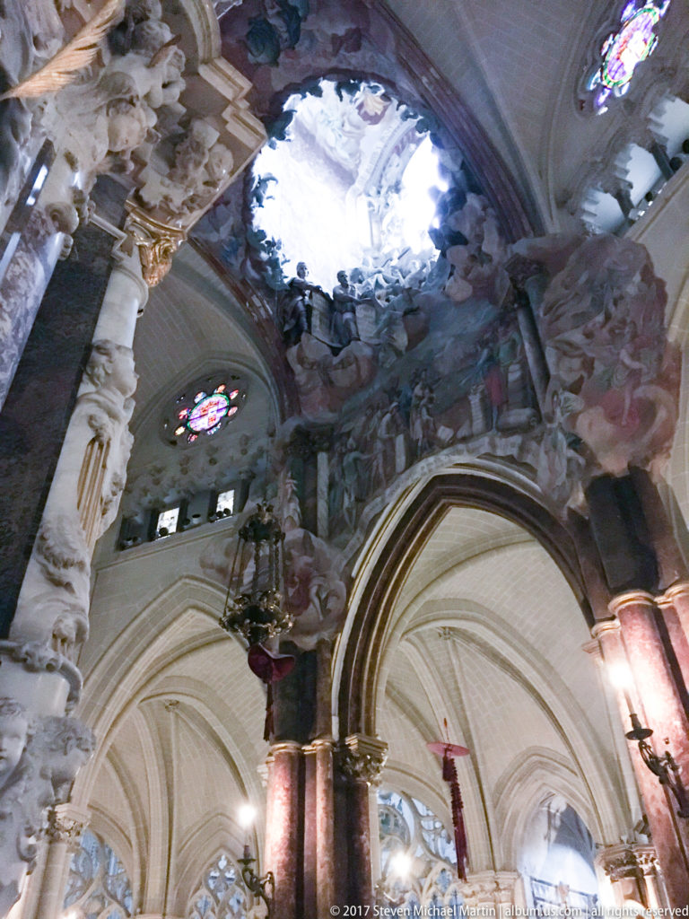 Upward view of a bright oculus surrounded by painted figures, clouds, sculpture, Gothic ribs, and stained glass.