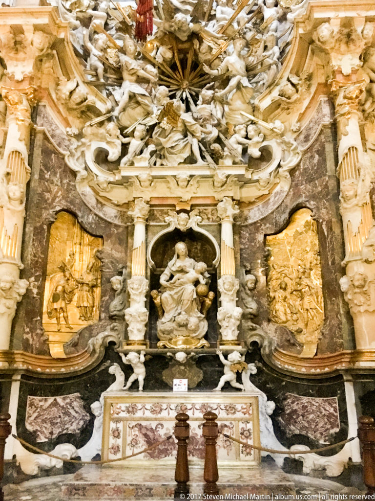 Frontal lower view of the Transparente with a central seated figure, gilded side reliefs, pale columns, white sculpture, and altar table.