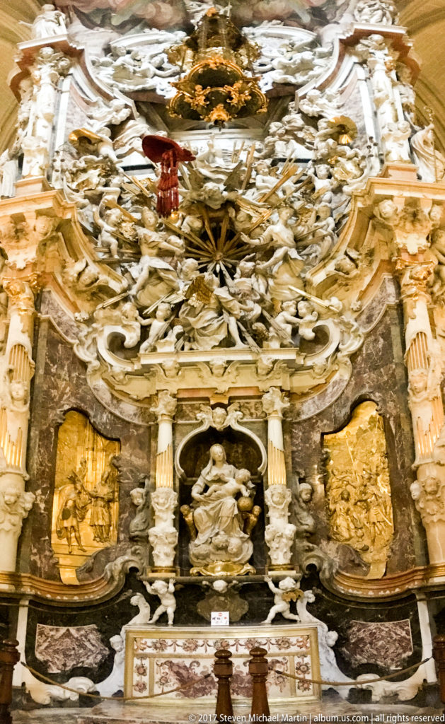 Lower frontal view of Toledo Cathedral’s Transparente with altar table, seated niche figure, gilded relief panels, columns, and sculpture above.