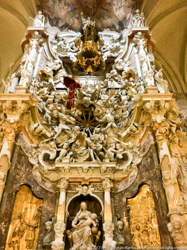 Frontal upward view of the Transparente with colored marble, seated figure, dense white sculpture, and a luminous painted opening above.