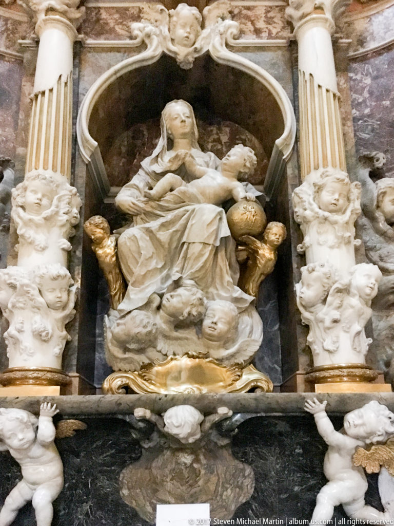 Seated robed stone figure in a dark niche between pale columns, surrounded by gilded accents and small sculpted figures.
