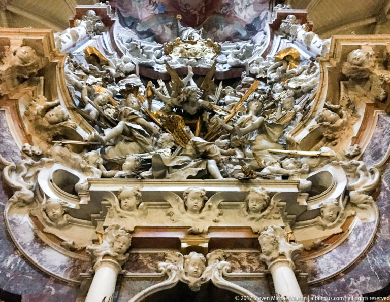 Dense Baroque cathedral sculpture with angels, clouds, columns, and an opening surrounded by painted and carved celestial forms.