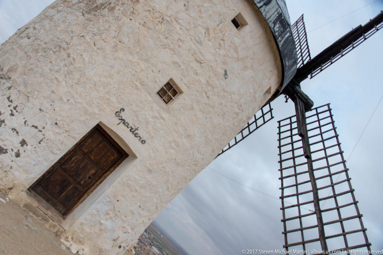 Tilted close view of a white windmill wall, wooden door, small window, dark roof, and lattice sail assembly.