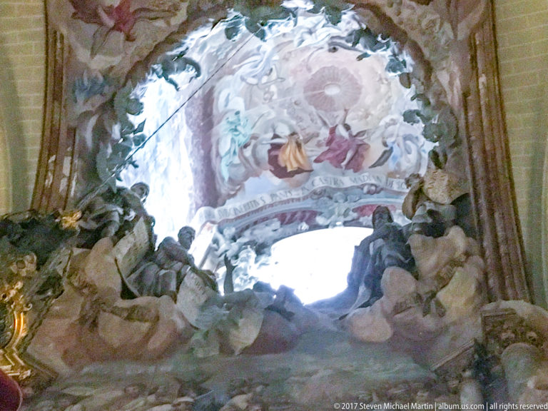 Close upward view of a bright cathedral opening surrounded by Baroque frescoes of figures and clouds with projecting pale sculpture.