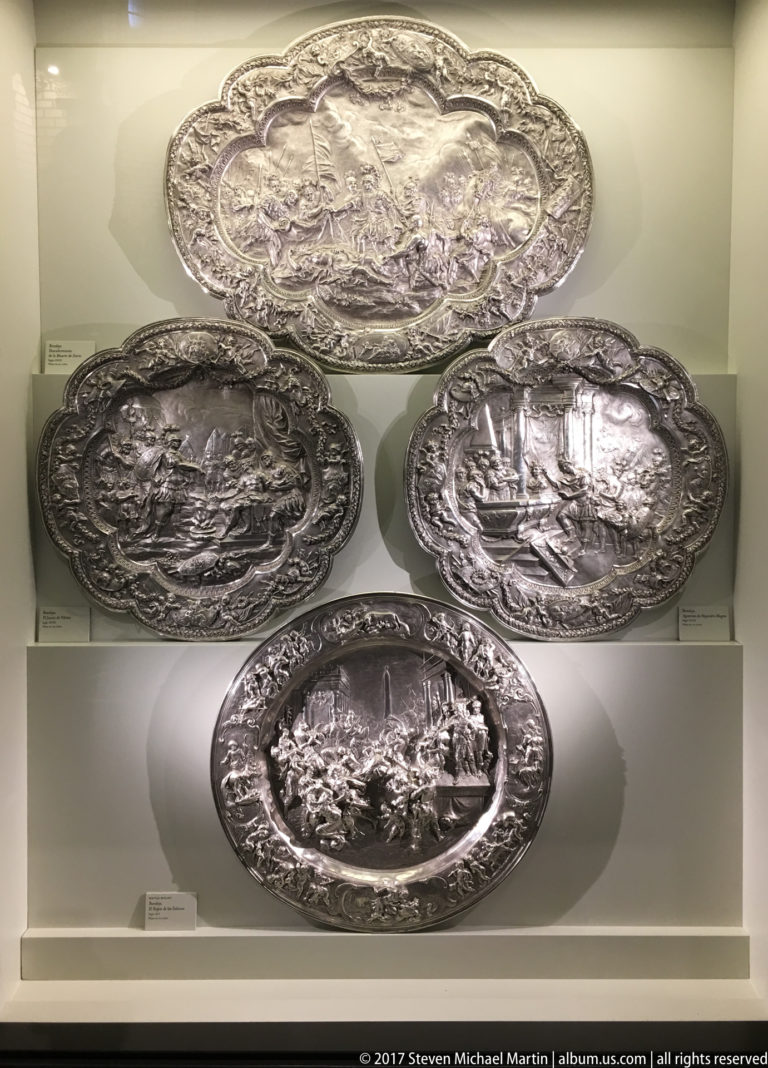 Four ornate silver platters with raised narrative reliefs displayed vertically on stepped museum mounts.
