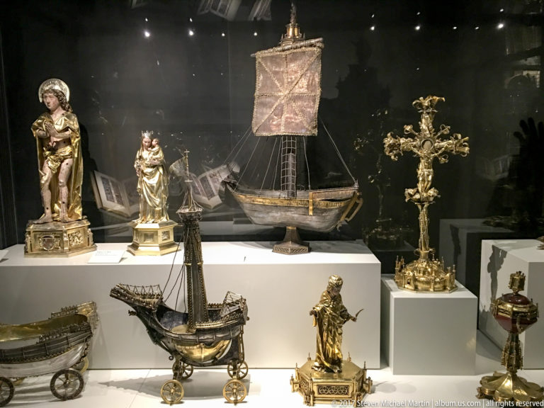 Museum display with sacred statuettes, an ornate cross, and several model sailing vessels arranged on white plinths.