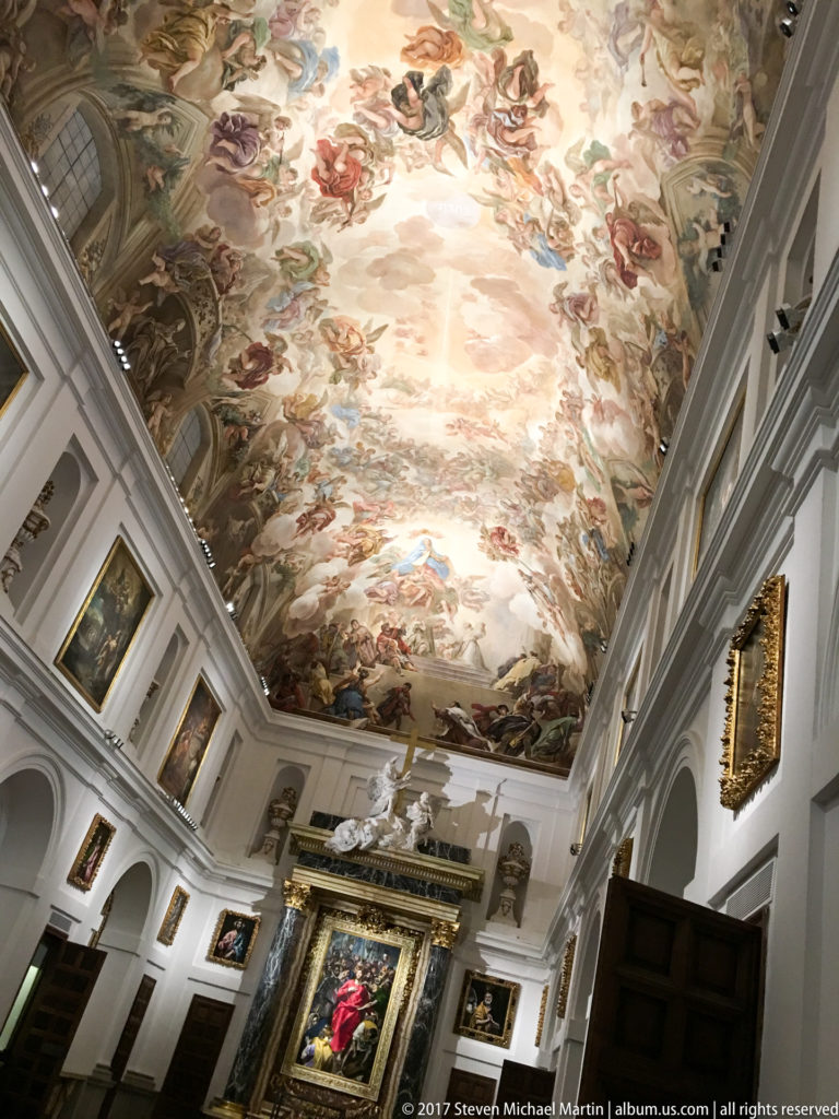 Long white church interior with a large frescoed ceiling, framed paintings along the walls, and an ornate devotional work at the far end.