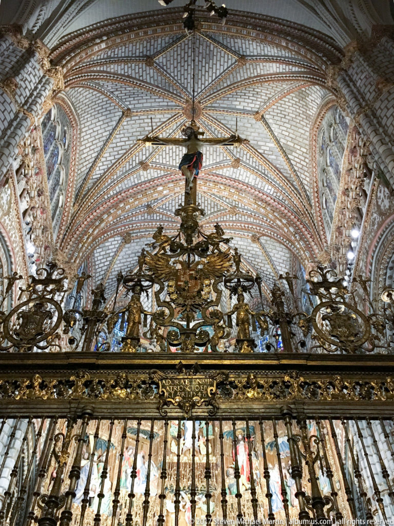 Upward view of an ornate gilded cathedral grille beneath patterned rib vaulting and stained-glass windows.