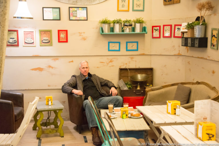 Man seated in a small Toledo café with colorful tins, potted plants, framed wall art, tables, and mismatched chairs.