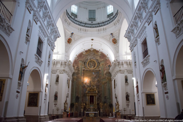 White Baroque church nave with deep side arches, balconies, a domed crossing, and an elaborate high altar.