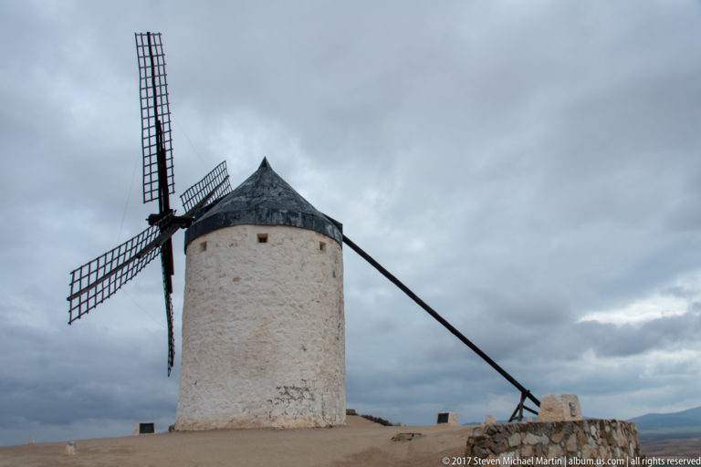 White cylindrical windmill with lattice sails, dark conical roof, and a long timber beam extending toward the ground.