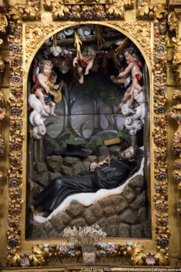 Reclining sacred figure in a rocky devotional scene beneath angels, enclosed by an ornate gilded architectural frame.