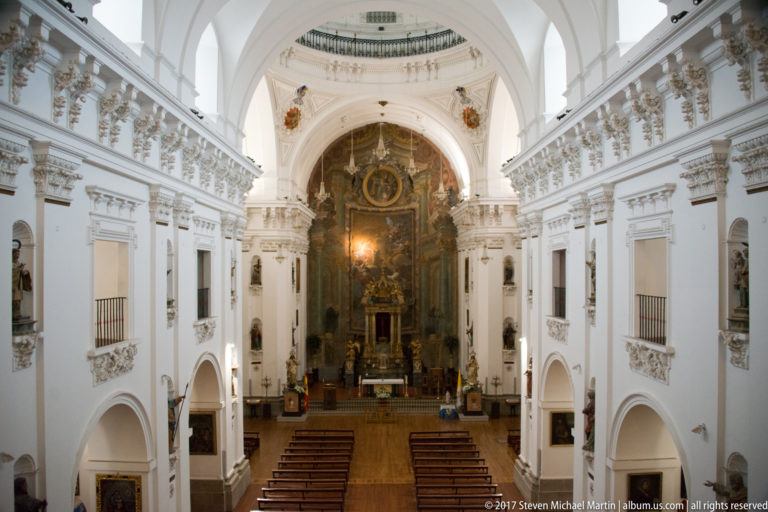 Elevated symmetrical view down a white Baroque church nave toward a dark monumental altar, with pews and side galleries.