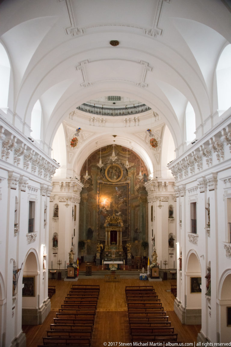 Wide frontal view of a white Baroque church interior with wooden pews, upper balustrades, vaulted ceiling, and dark high altar.