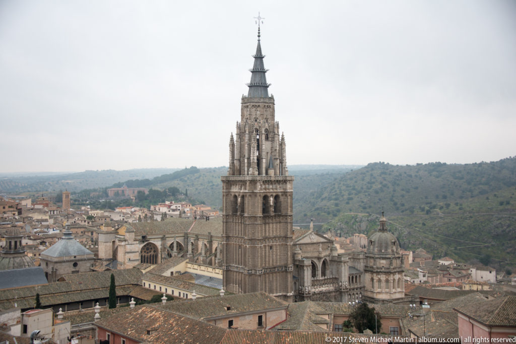 Toledo Cathedral’s Gothic tower rising above clustered historic rooftops, domes, and chapels with wooded hills beyond.