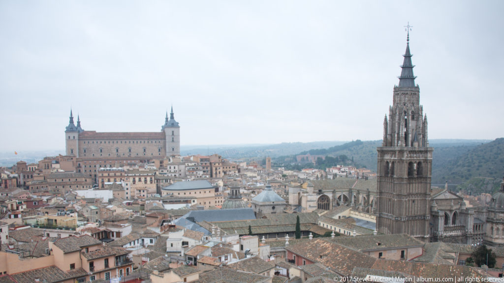 Panoramic Toledo skyline with the cathedral tower at right and the four-towered Alcázar rising on the distant ridge.