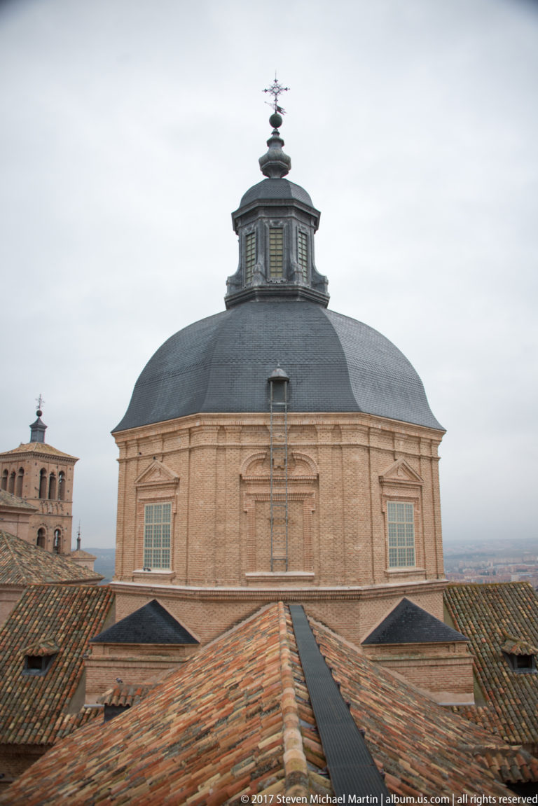 Large dark dome with polygonal drum and lantern rising above tiled roofs in the Toledo Cathedral complex.