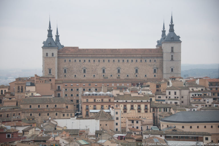 Telephoto view of the Alcázar of Toledo with four corner towers rising above dense historic buildings.