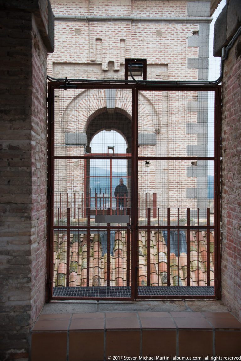 Metal gate blocking a narrow passage between brick and stone walls, with tiled cathedral roofs visible beyond.