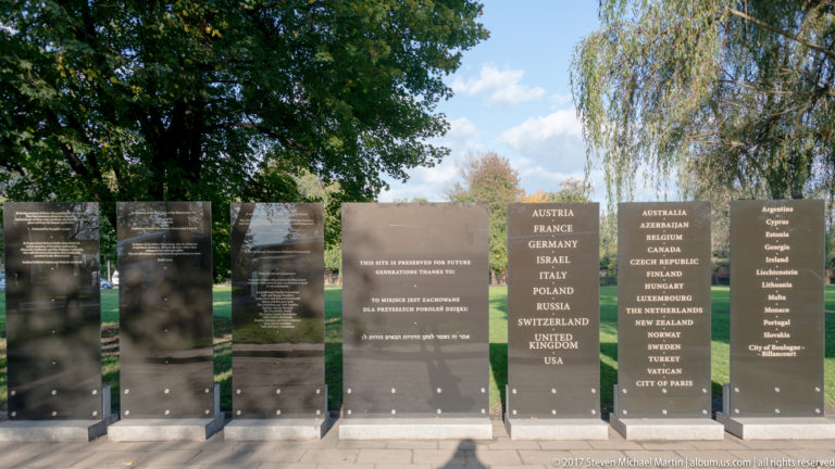 Reflective memorial plaques with multilingual inscriptions and lists of countries stand outdoors among trees at Auschwitz.