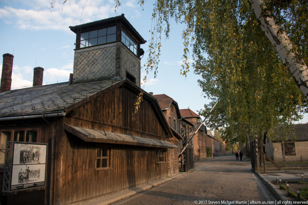 Elevated enclosed guard tower rises above a wooden camp building beside an internal lane lined with brick blocks.