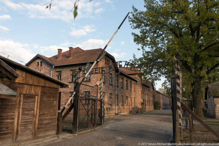 Oblique view through the Auschwitz I entrance beneath the iron “Arbeit macht frei” sign and raised barrier.