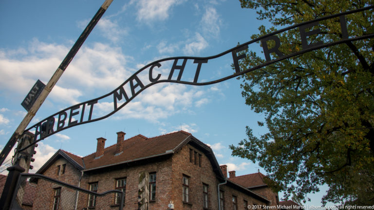 Black iron “Arbeit macht frei” sign arches above the Auschwitz I entrance, with brick buildings below.