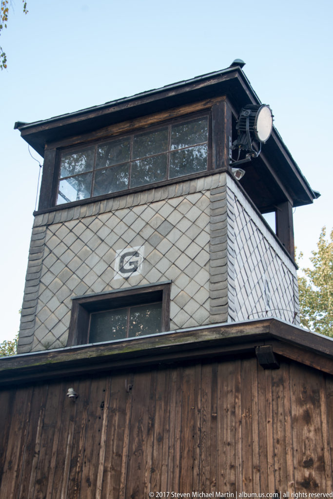 Low-angle close view of a windowed guard tower with shingled upper walls, timber base and mounted lamp.