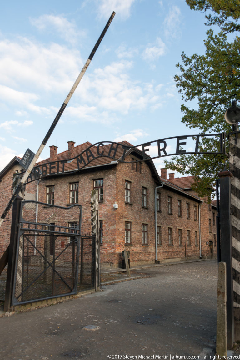 Wide frontal view of the Auschwitz I entrance with “Arbeit macht frei,” raised barrier, gate and brick block.