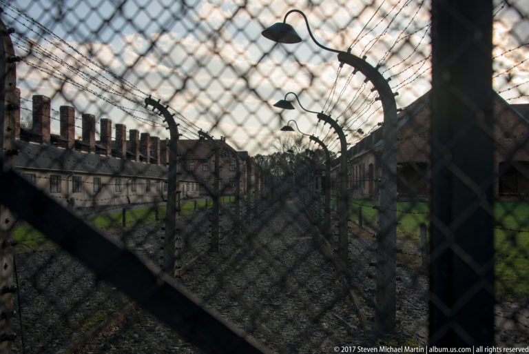 Layered mesh and barbed-wire fencing on curved concrete posts runs between long barracks at Auschwitz I.