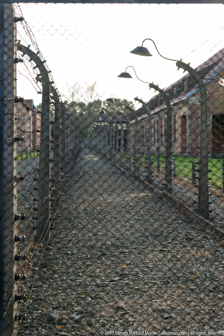 Gravel corridor enclosed by parallel barbed-wire fences on curved concrete posts, with camp buildings beyond.