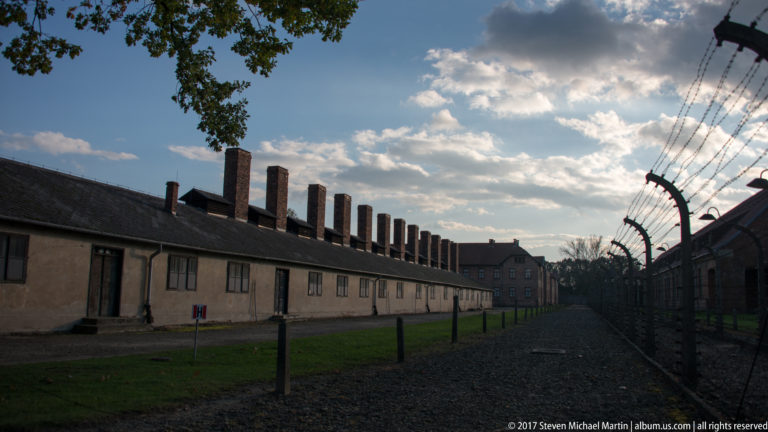 Long pale barrack with many chimneys runs beside a barbed-wire fence under a backlit sky.