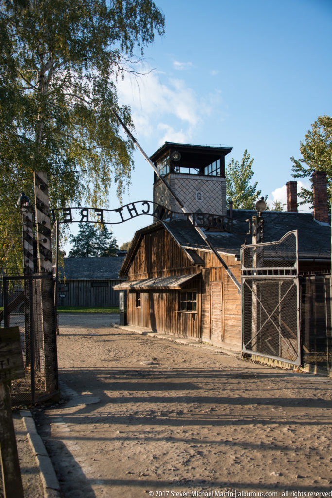 Interior-facing view of Auschwitz I entrance with reversed “Arbeit macht frei” lettering, open gates and guard tower.
