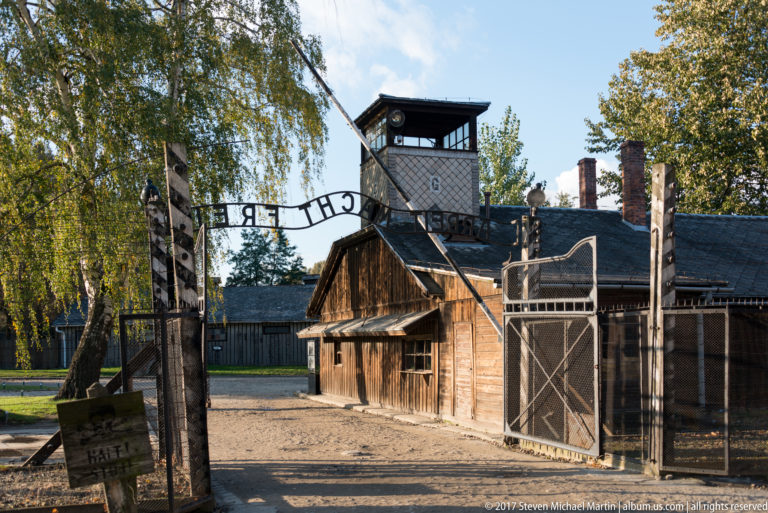 Open Auschwitz I gate seen from inside, with reversed entrance lettering, wooden building and guard tower.