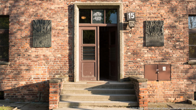 Brick entrance to Auschwitz I Block 15 with number plaque, brown doorway, steps and dark wall plaques.
