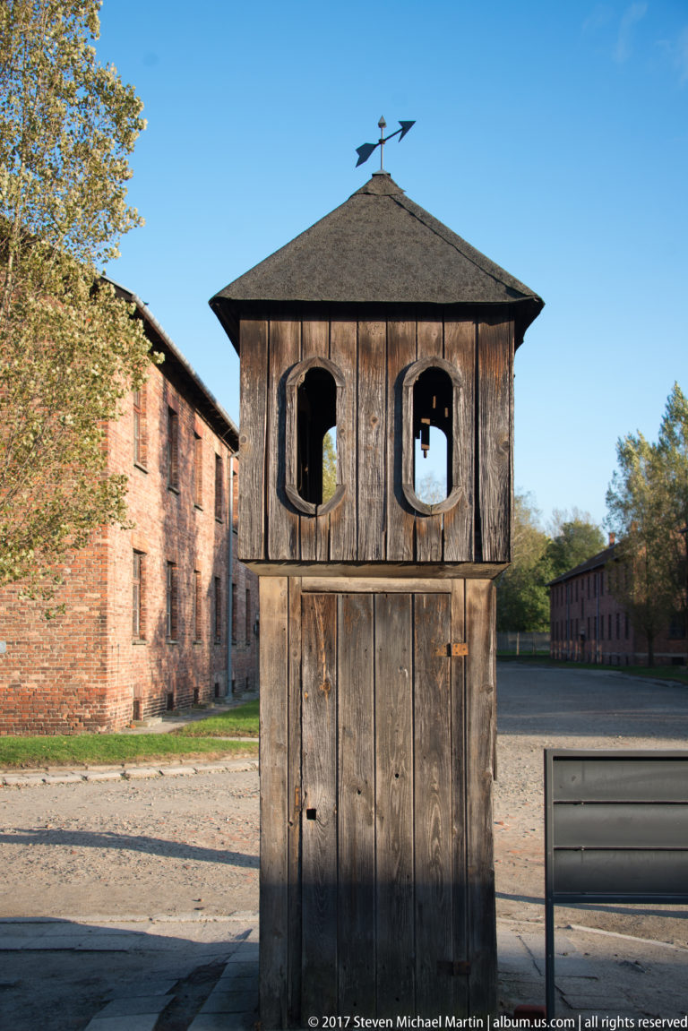 Dark wooden gallows structure with two upper openings and a hipped roof stands between brick blocks.