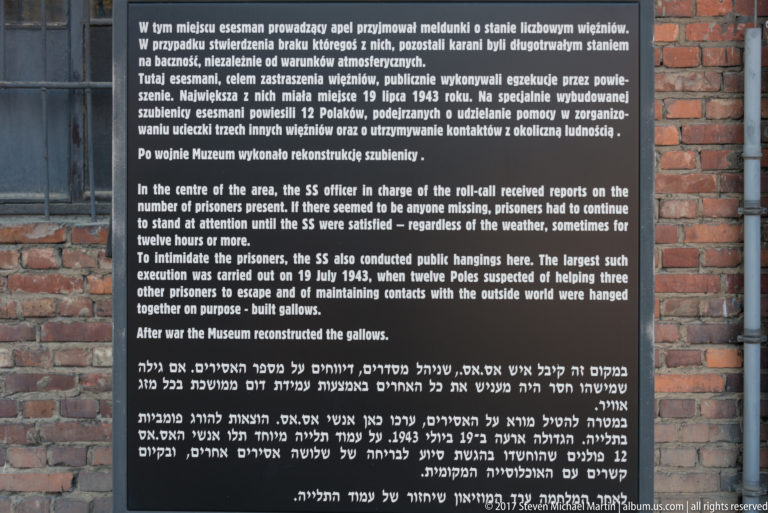 Black multilingual panel on a brick wall explains the gallows, roll calls and public executions at Auschwitz I.