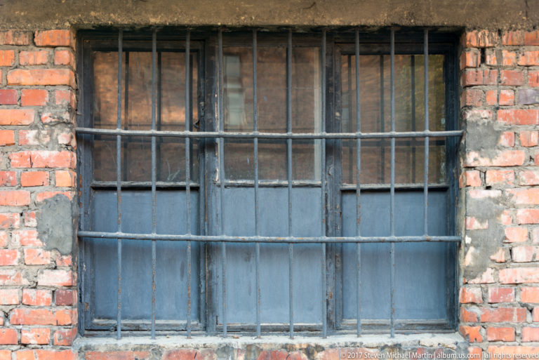 Close view of a large multi-pane window secured by heavy iron bars in a weathered red-brick wall.