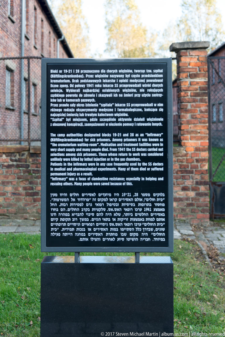 Freestanding black multilingual historical panel at Auschwitz I, with red-brick camp blocks behind it.