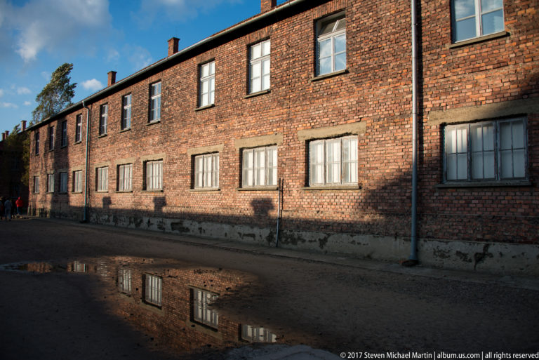 Long two-story red-brick Auschwitz I block with repeated windows reflected partly in a foreground puddle.