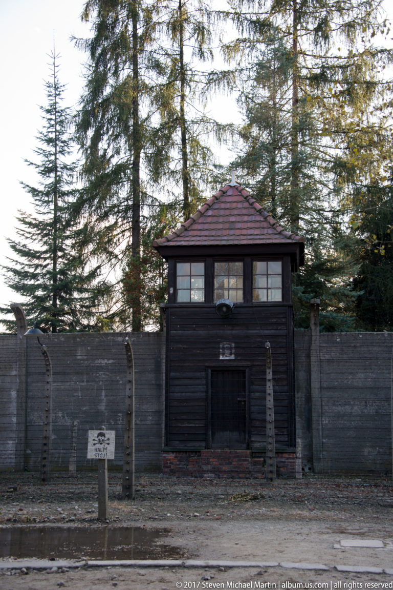 Dark wooden guard tower rises behind a concrete wall and barbed-wire perimeter fence among tall evergreen trees.