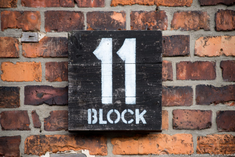 Black wooden sign reading “11 BLOCK” in white letters mounted on a weathered red-brick wall.