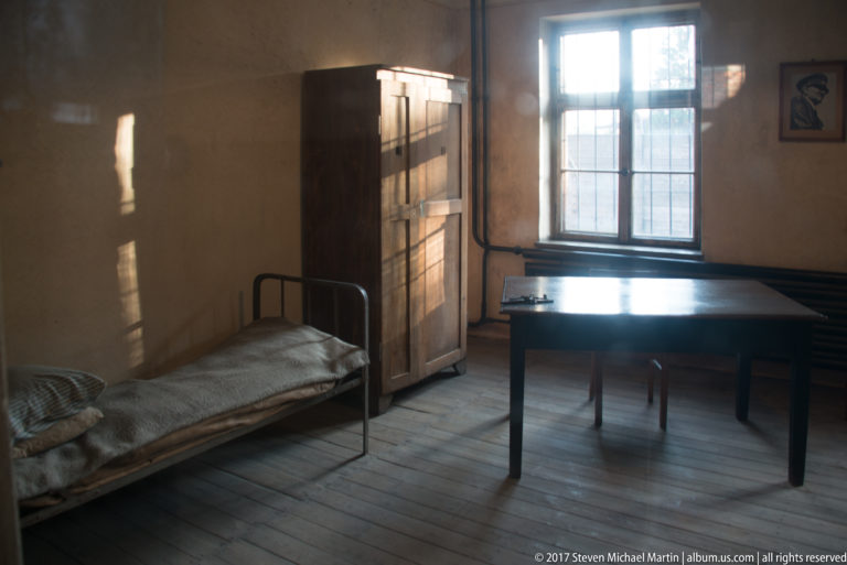 Plain Block 11 room with narrow metal bed, wooden table, cupboard, benches and daylight through a multi-pane window.