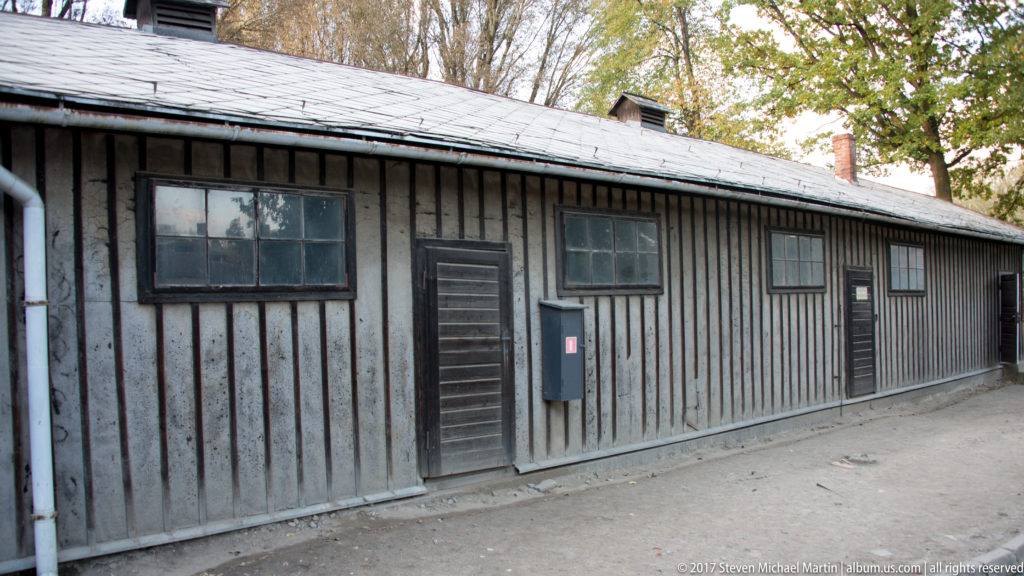 Long weathered wooden barrack with vertical boards, small windows and plain doors beside a gravel path.