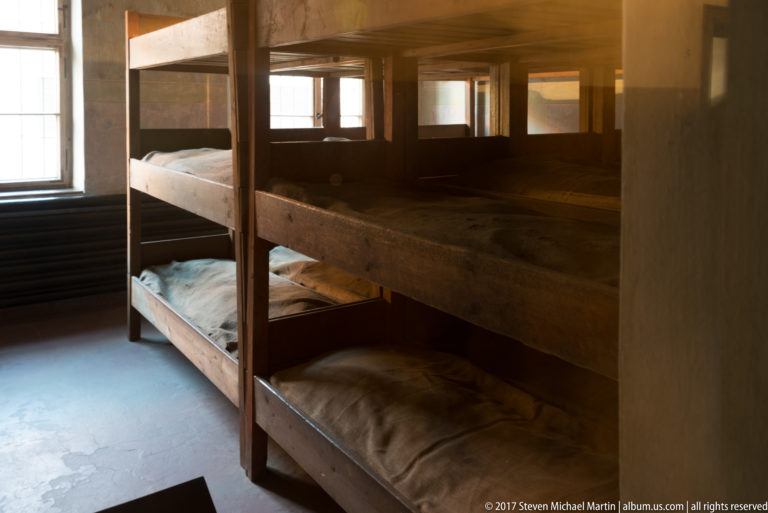 Heavy wooden bunk beds with thin bedding fill a dim Block 11 room lit by windows at left and rear.