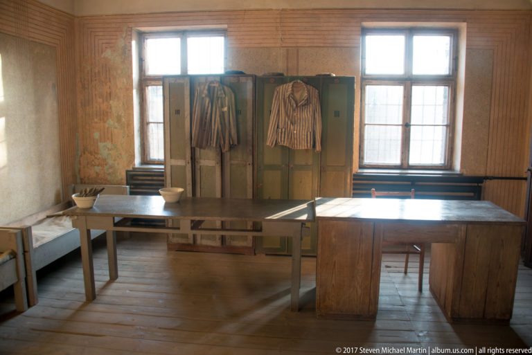 Striped prisoner garments hang from wooden lockers behind plain tables, stools and bowls in a worn room.