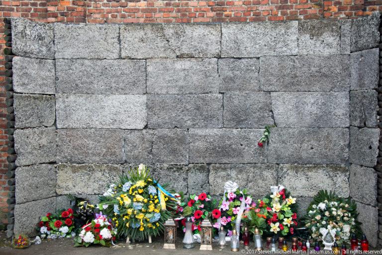 Flowers and wreaths rest against the gray reconstructed Execution Wall in a brick courtyard at Auschwitz I.