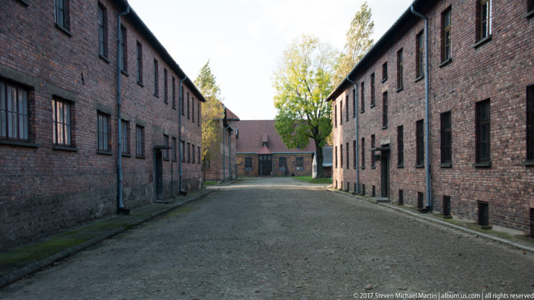 Straight gravel lane between two long red-brick Auschwitz I blocks, with trees and a smaller building ahead.