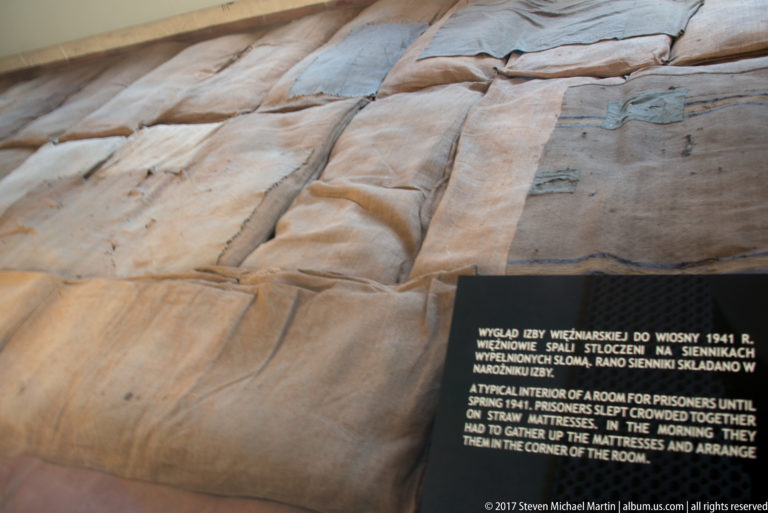 Large tan straw mattresses lie piled together beside an interpretive label describing early prisoner sleeping conditions.
