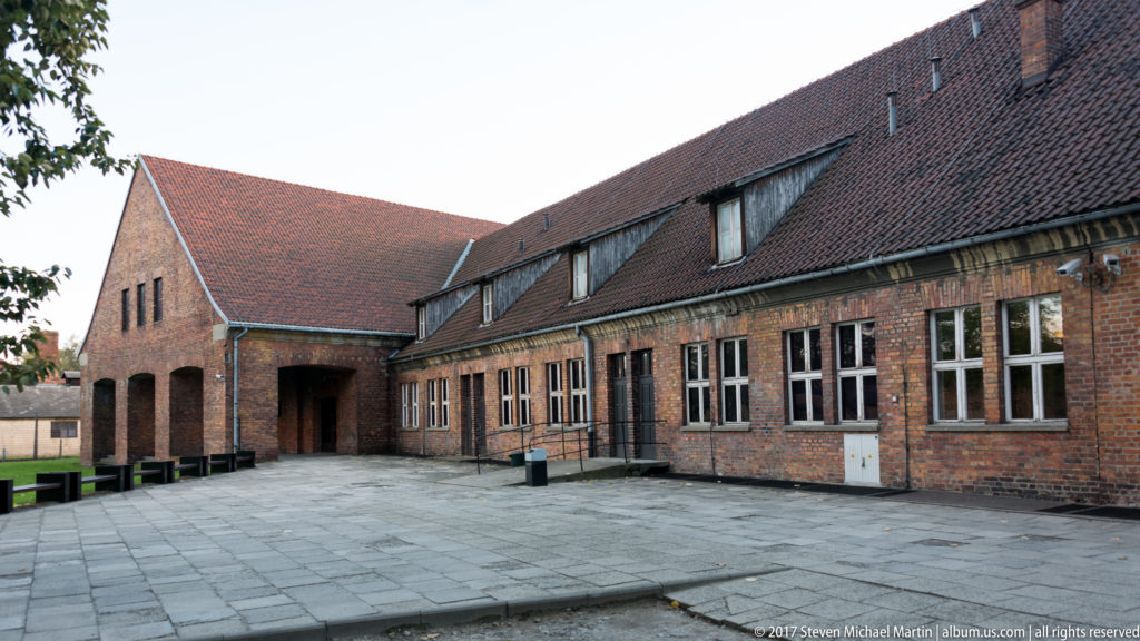 Long red-brick building with tiled roof, rows of windows and a deep passage beside a paved courtyard.